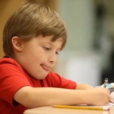 Young student in a red shirt writing at a desk in a classroom