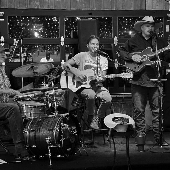 Black-and-white photo of three members of the band Cross Country performing on stage, with a drummer at left, a seated guitarist at center and a standing guitarist in a cowboy hat at right