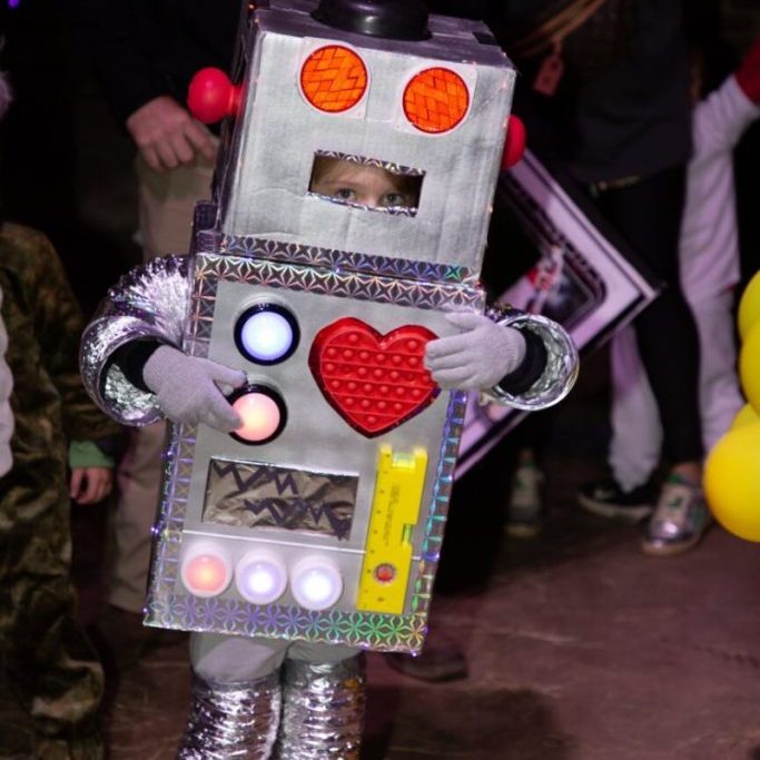 Child in a homemade robot costume with a red heart on the chest at a nighttime Halloween event