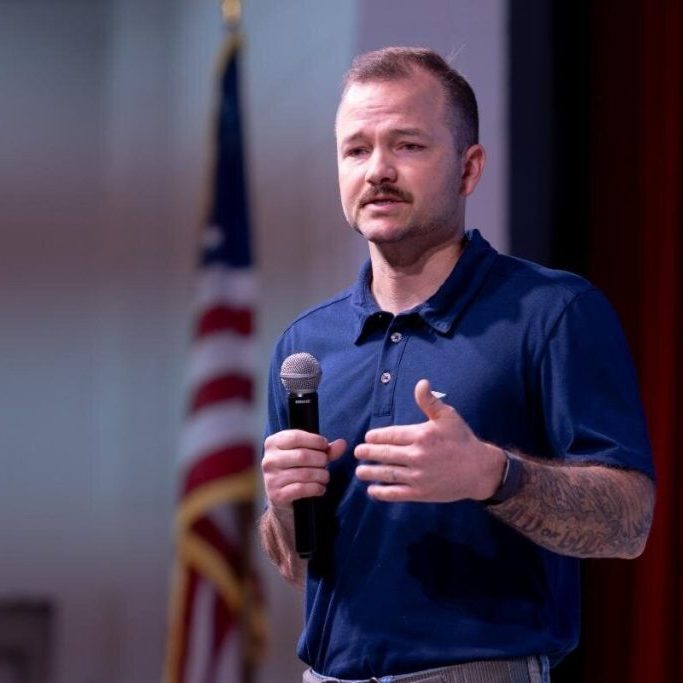 A man speaking into a microphone on stage in front of an American flag